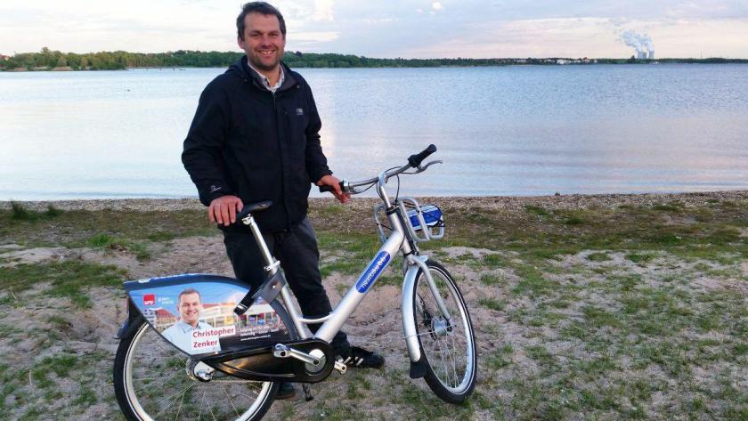 Der Fraktionsvorsitzende der Leipziger SPD, Christopher Zenker, lächelnd mit seinem Wahlkampffahrad am Cospudener See