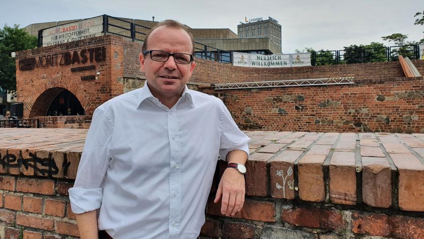 René Hobusch lehnt an der Moritzbastei. Er trägt ein weißes Hemd und eine graue Anzughose.